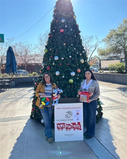 Two women stand by a holiday tree holding toys next to a Toys for Tots donation box outdoors.