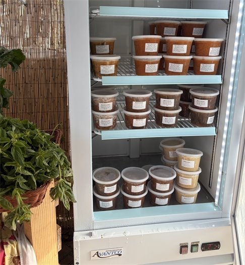 Containers of house-made soups are stacked in a small refrigerator case at The Coop, with fresh basil visible beside it.